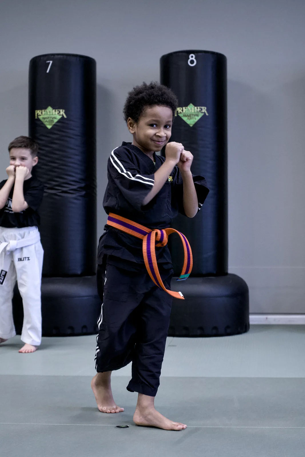 A small child in an orange belt standing on one leg beside a punch bag