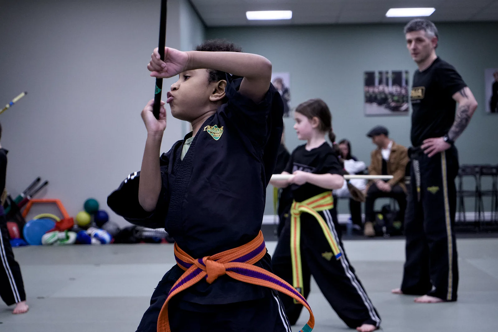 A child mid-strike with a padded weapon, the instructor watching