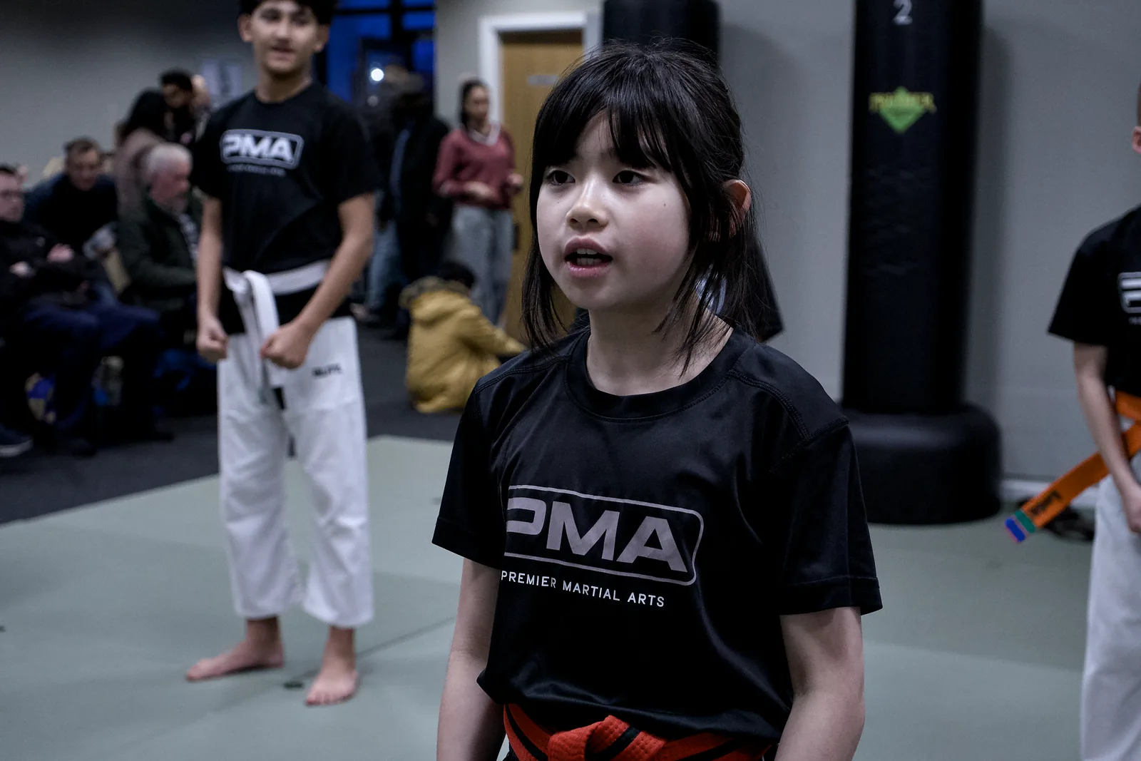 A girl in a PMA shirt and orange belt standing ready, watching the instructor