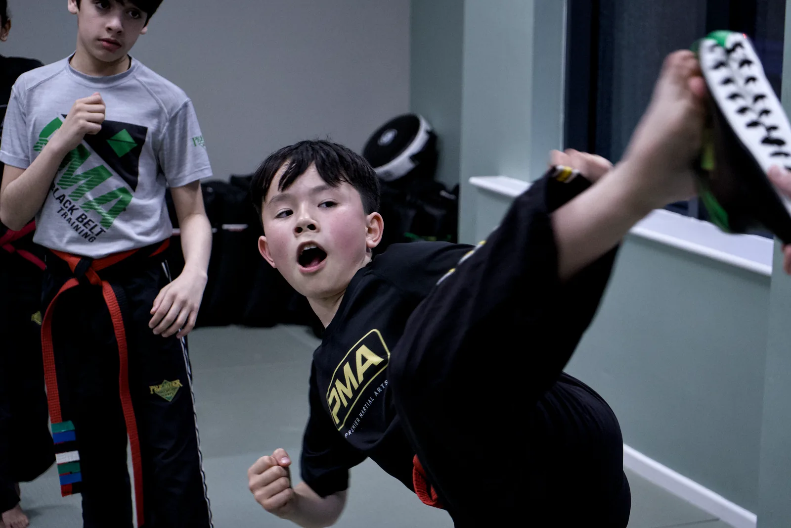 A young boy in a PMA uniform throwing a high kick, mouth open with effort