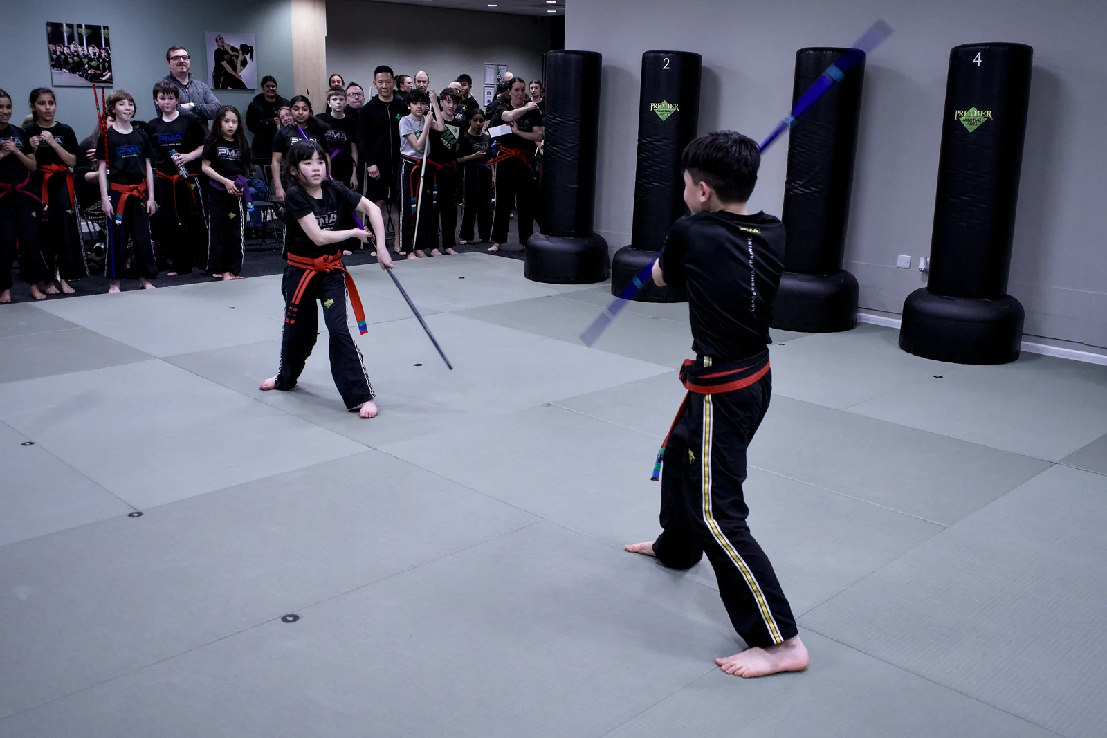 Two children facing each other across the mat holding training sticks