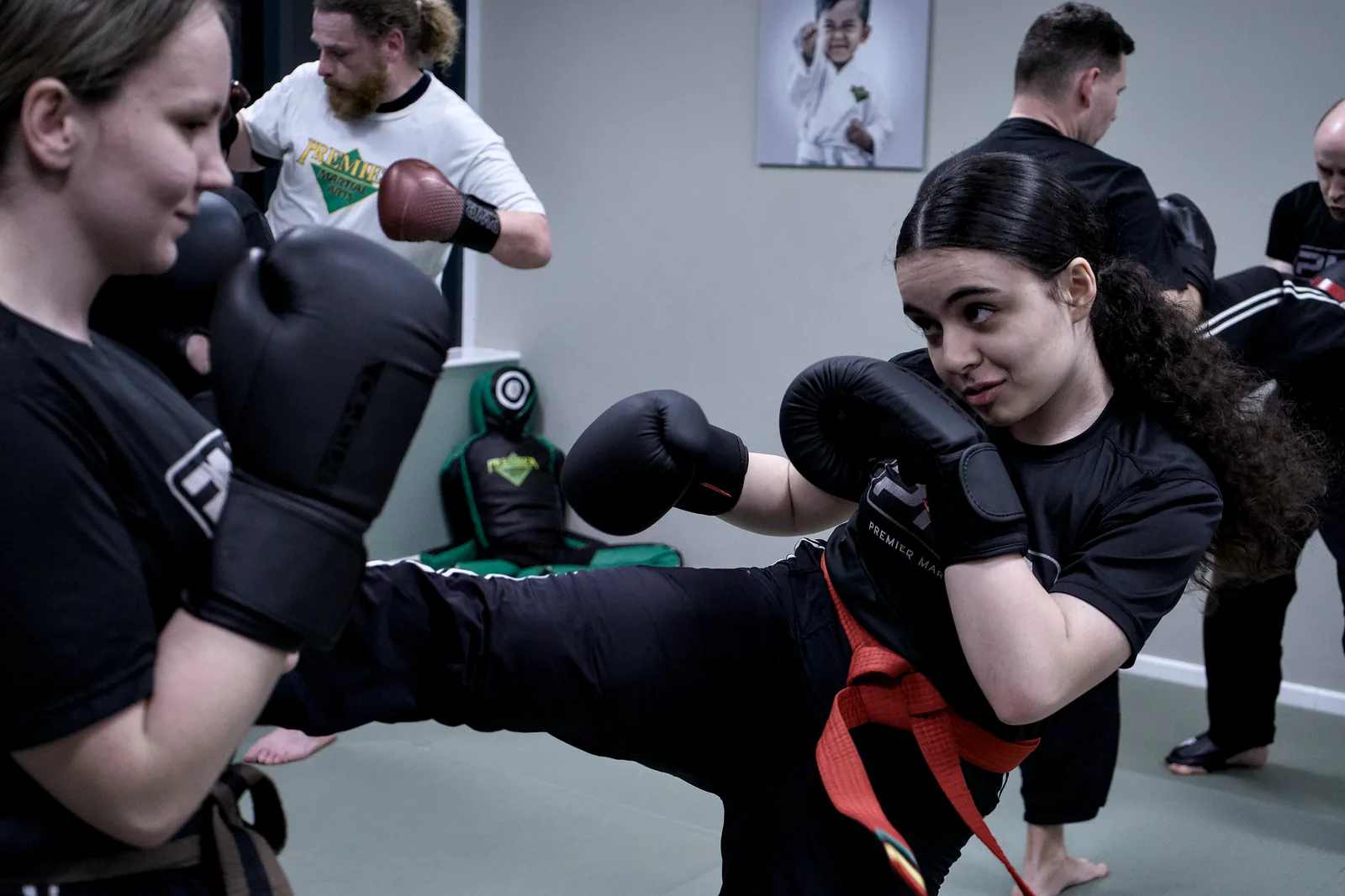 An adult student landing a kick on a pad, another student holding it steady